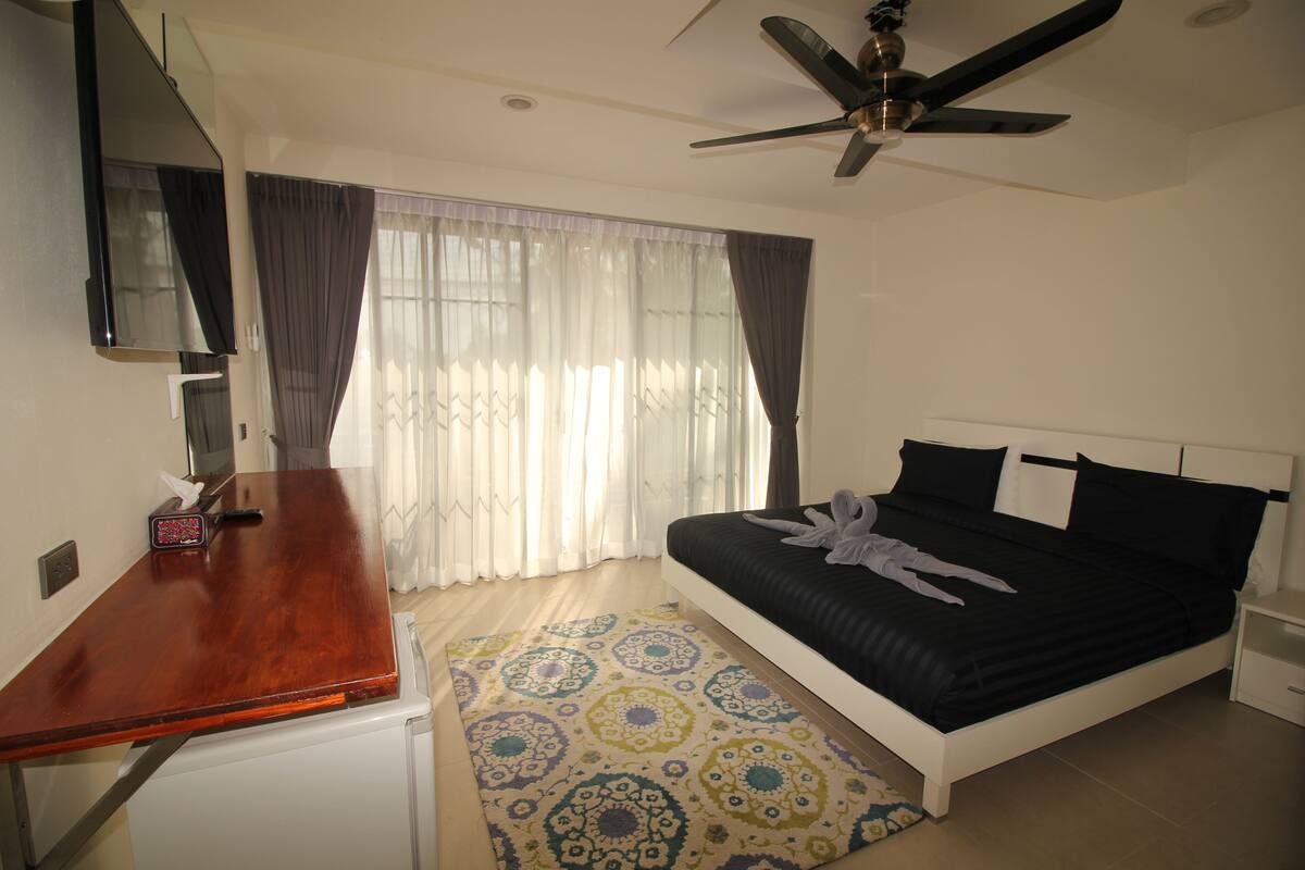 Une chambre à coucher avec un lit, un bureau, un ventilateur de plafond et une fenêtre avec des rideaux.