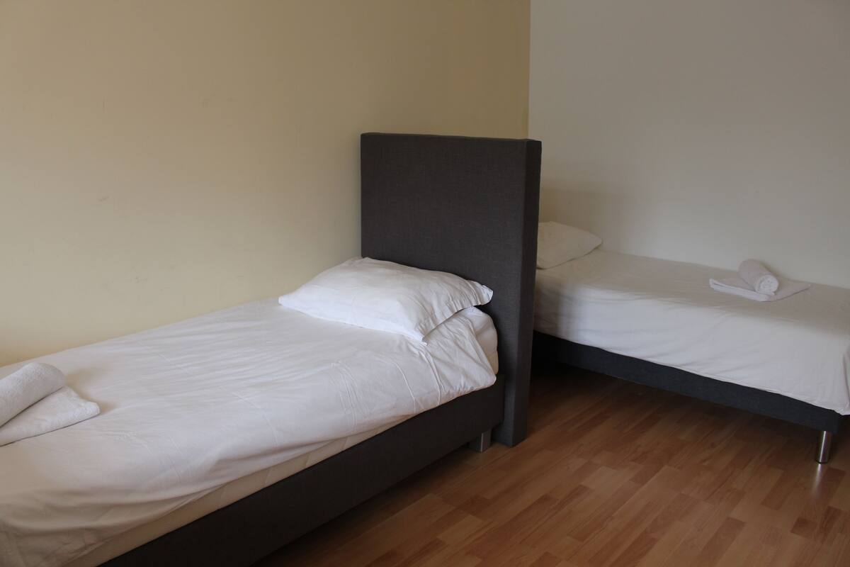 Two single beds with white linens and pillows, a wooden floor, and a plain wall.