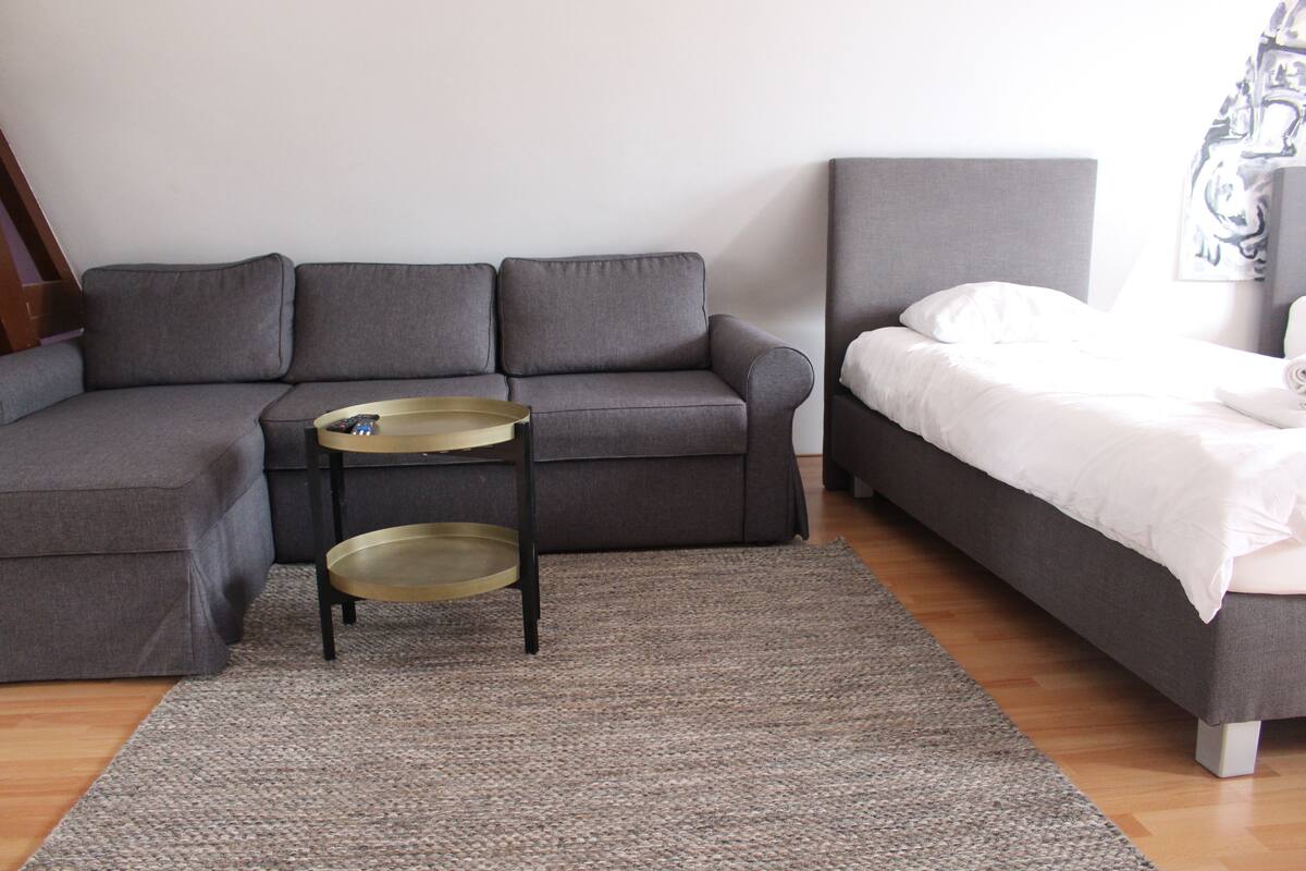 A modern living room with a grey sofa, a small round table, and a bed with white bedding.