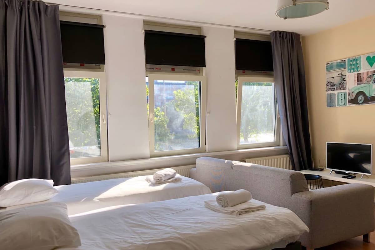 A hotel room with two beds, a sofa, a TV, and large windows with curtains.