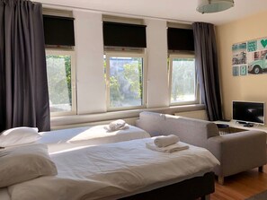 Une chambre d’hôtel avec deux lits, un canapé, une télévision et de grandes fenêtres équipées de rideaux.
