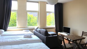 A hotel room with two beds, a desk with a computer, and a view of trees through large windows. - MyCityLofts - Aelbrechtskade (Rotterdam)