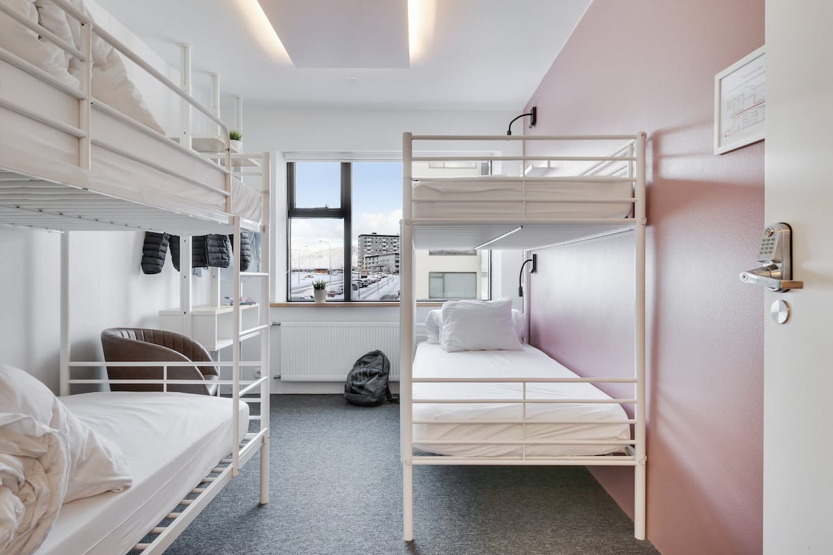 Ein Schlafsaalzimmer mit Etagenbetten, einem Fenster mit Blick auf Gebäude, einem Heizkörper und einer Tür mit Schloss.