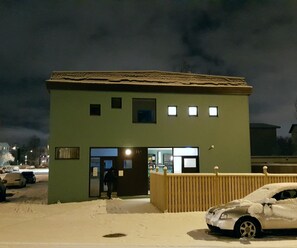 Um pequeno edifício térreo com telhado plano e fachada verde. Há uma cerca de madeira na frente, e um carro coberto de neve à direita.