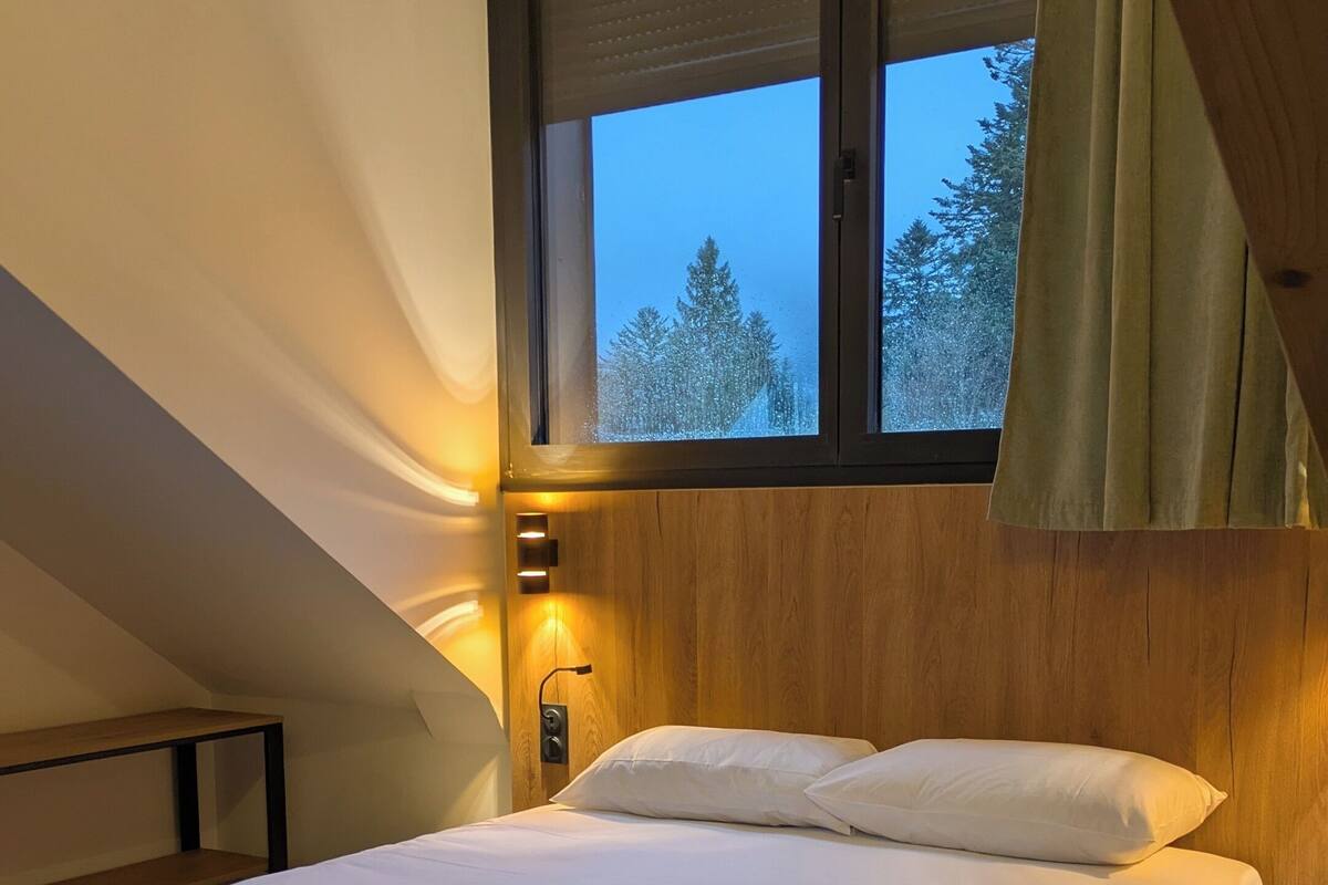 A bedroom with a bed, a wooden headboard, a window with a view of trees, and a wall-mounted light.