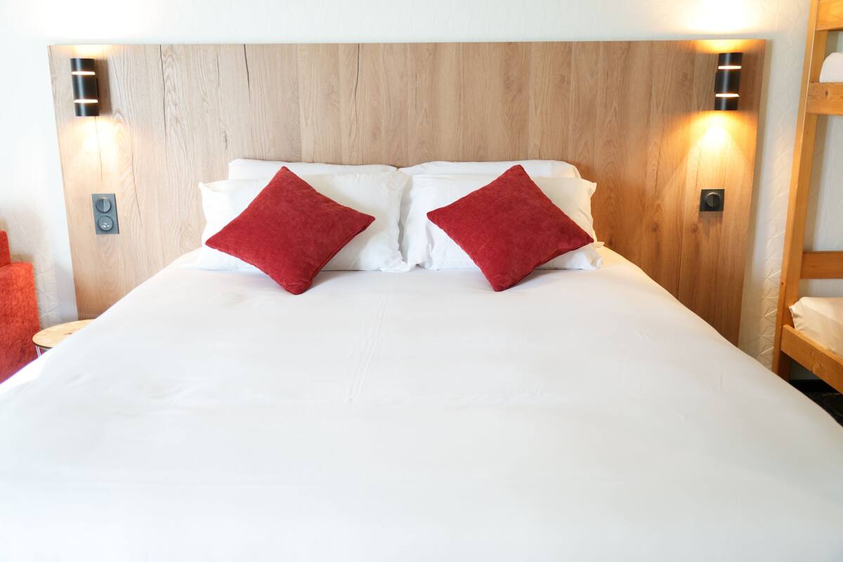 A large bed with white linens and two red pillows, a wooden headboard, and wall-mounted lighting.