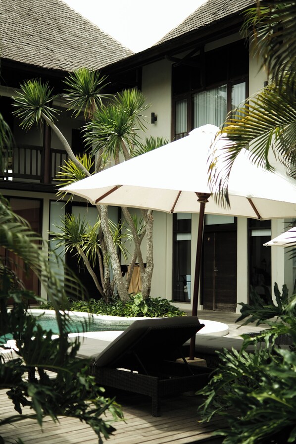 Uma área à beira da piscina com espreguiçadeira, guarda-sol e plantas tropicais.