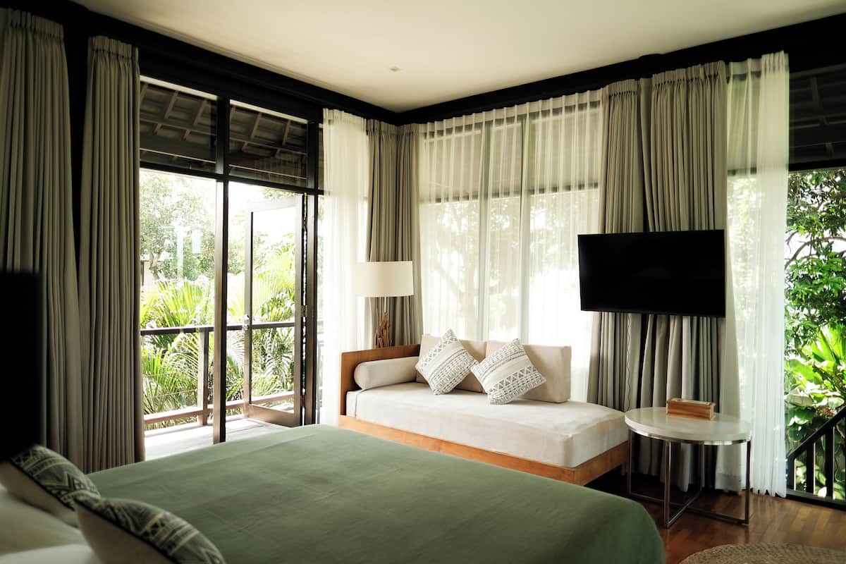 A hotel room with a large bed, a TV, a small table, and a view of greenery outside.