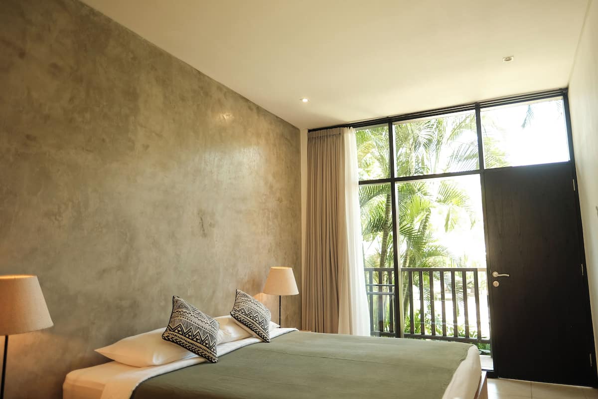 A bedroom with a large bed, two bedside lamps, a view of palm trees through a window, and a dark door.