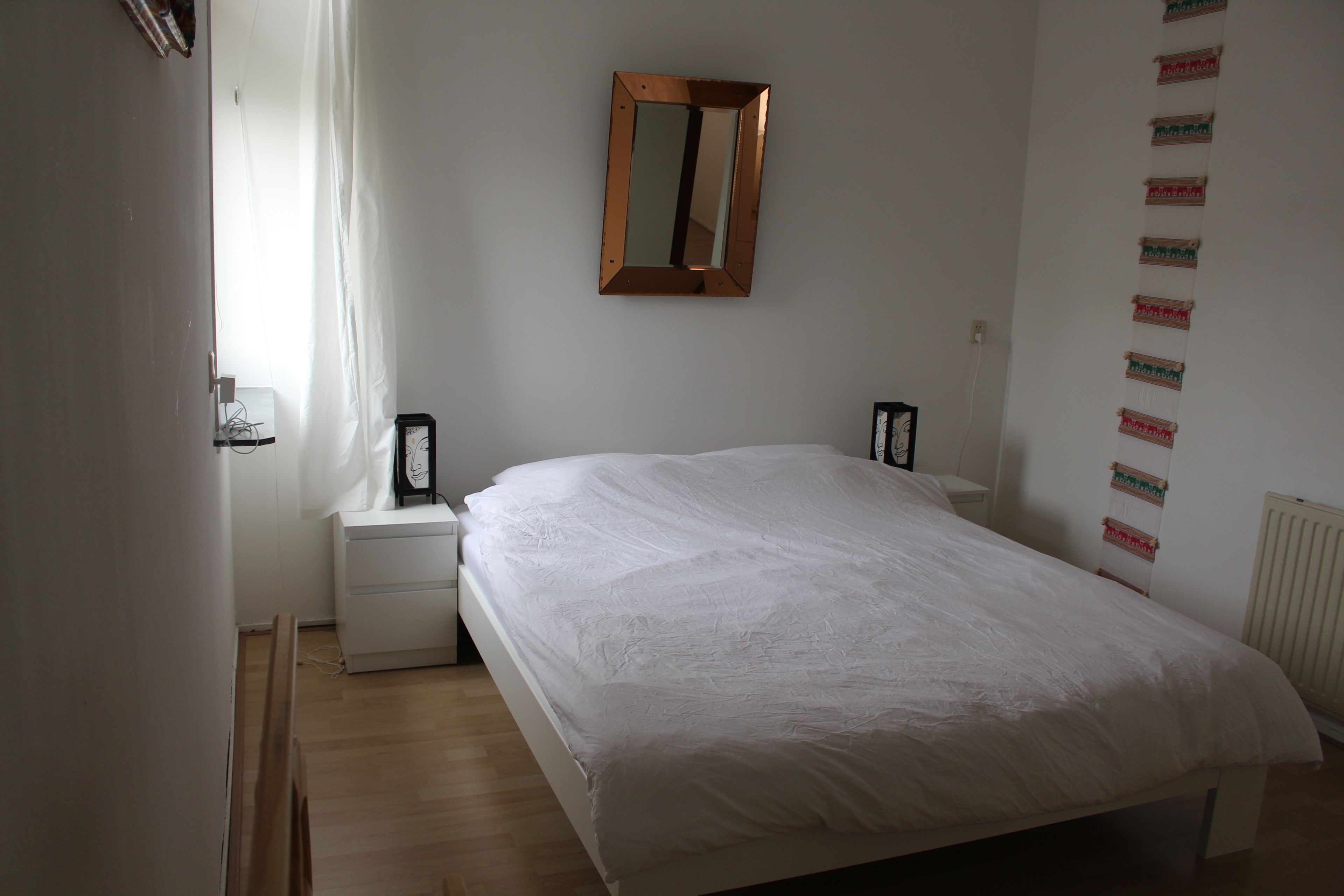 A bedroom with a large bed, a nightstand, a wall-mounted mirror, and a window with curtains.