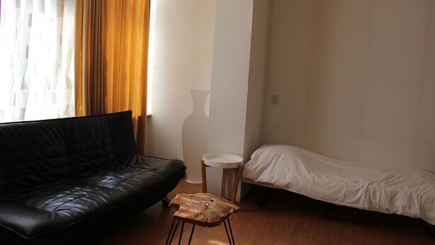 A room with a black leather sofa, a small wooden stool, a white bedside table, and a single bed. - MyCityLofts - Schiekade (Rotterdam)