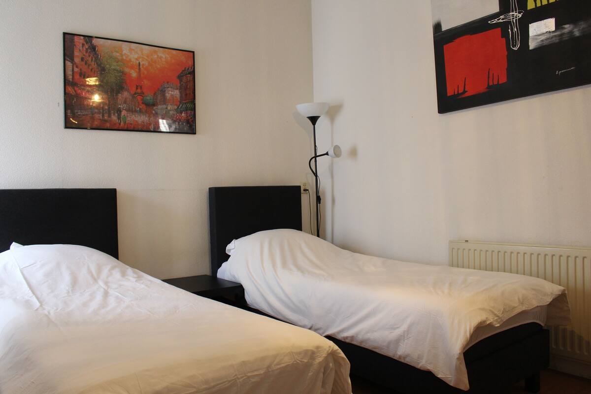A hotel room with two single beds, a nightstand, a lamp, and a painting on the wall.