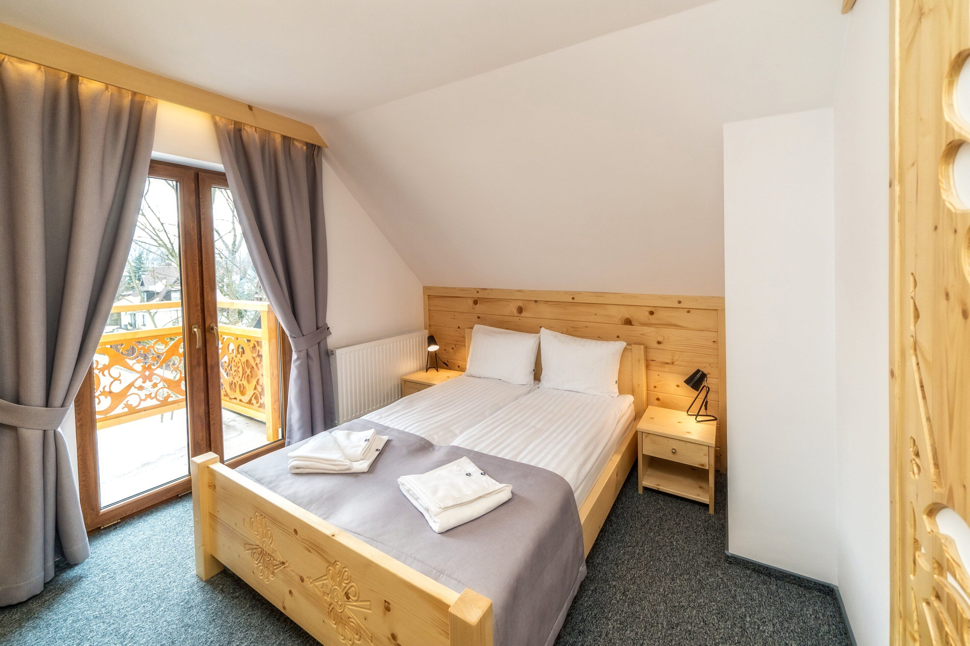 Un dormitorio con cabecera de madera, cama doble con ropa blanca, mesita de noche, lámpara y vistas a un balcón con cortinas.