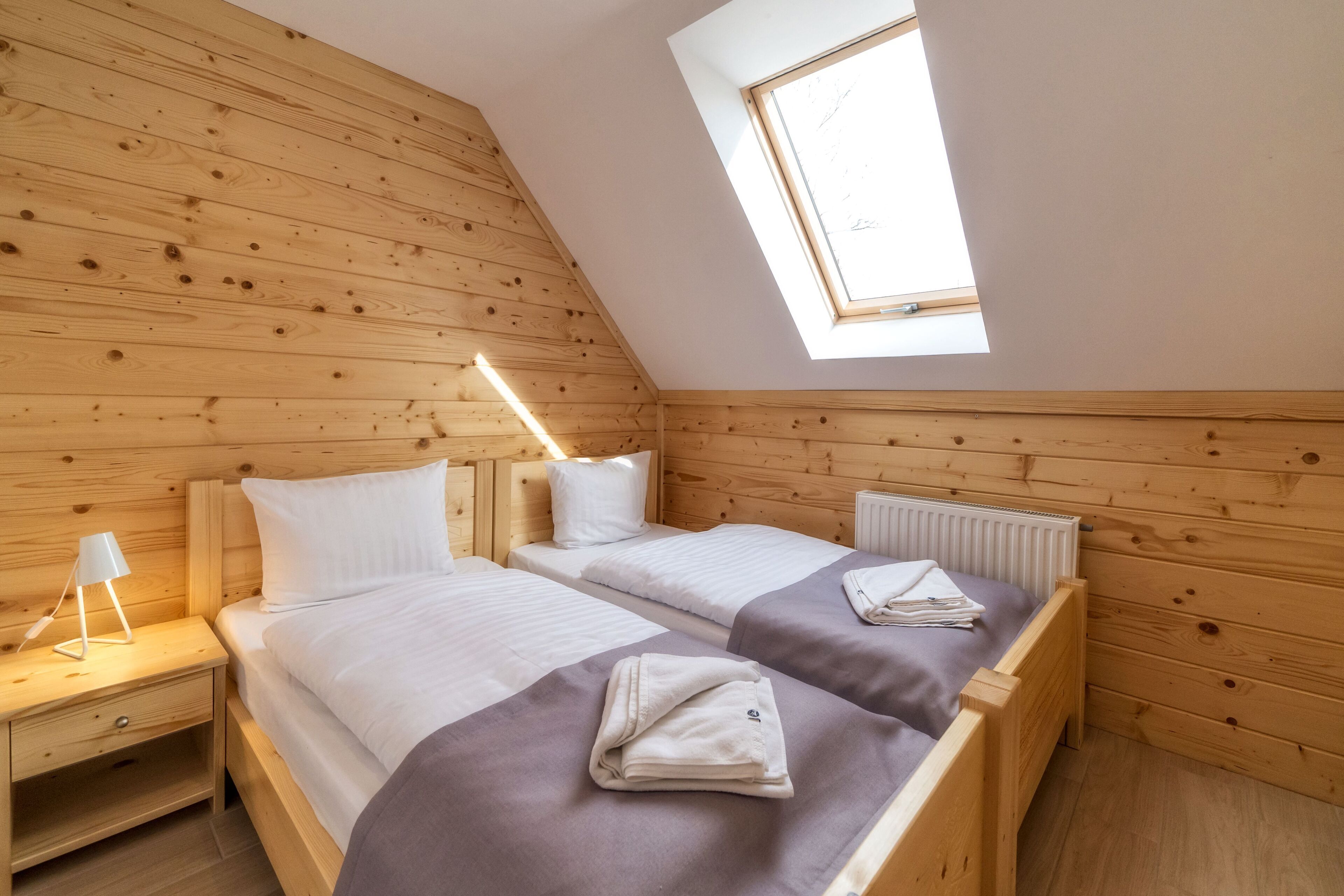 A room with two wooden beds, a skylight, a nightstand with a lamp, and a radiator.