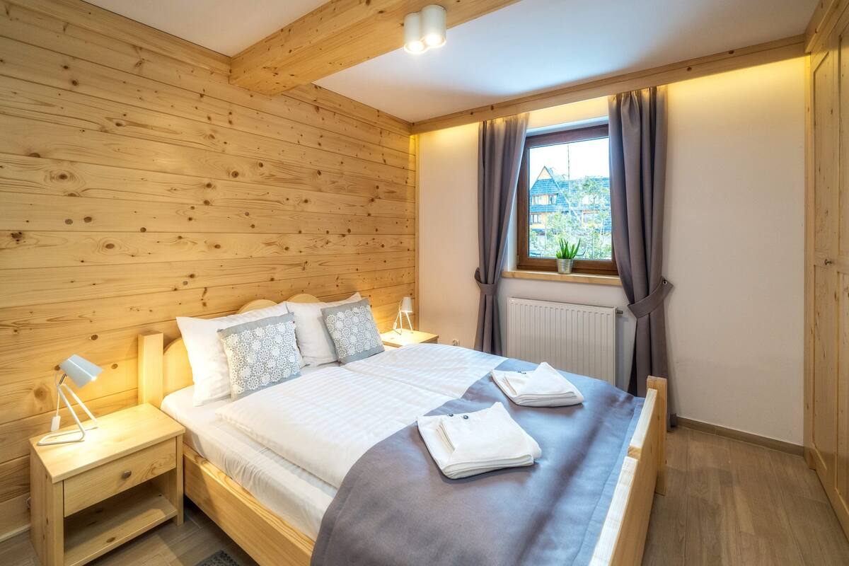 A wooden-paneled bedroom with a bed, nightstands, a window with curtains, and a radiator.