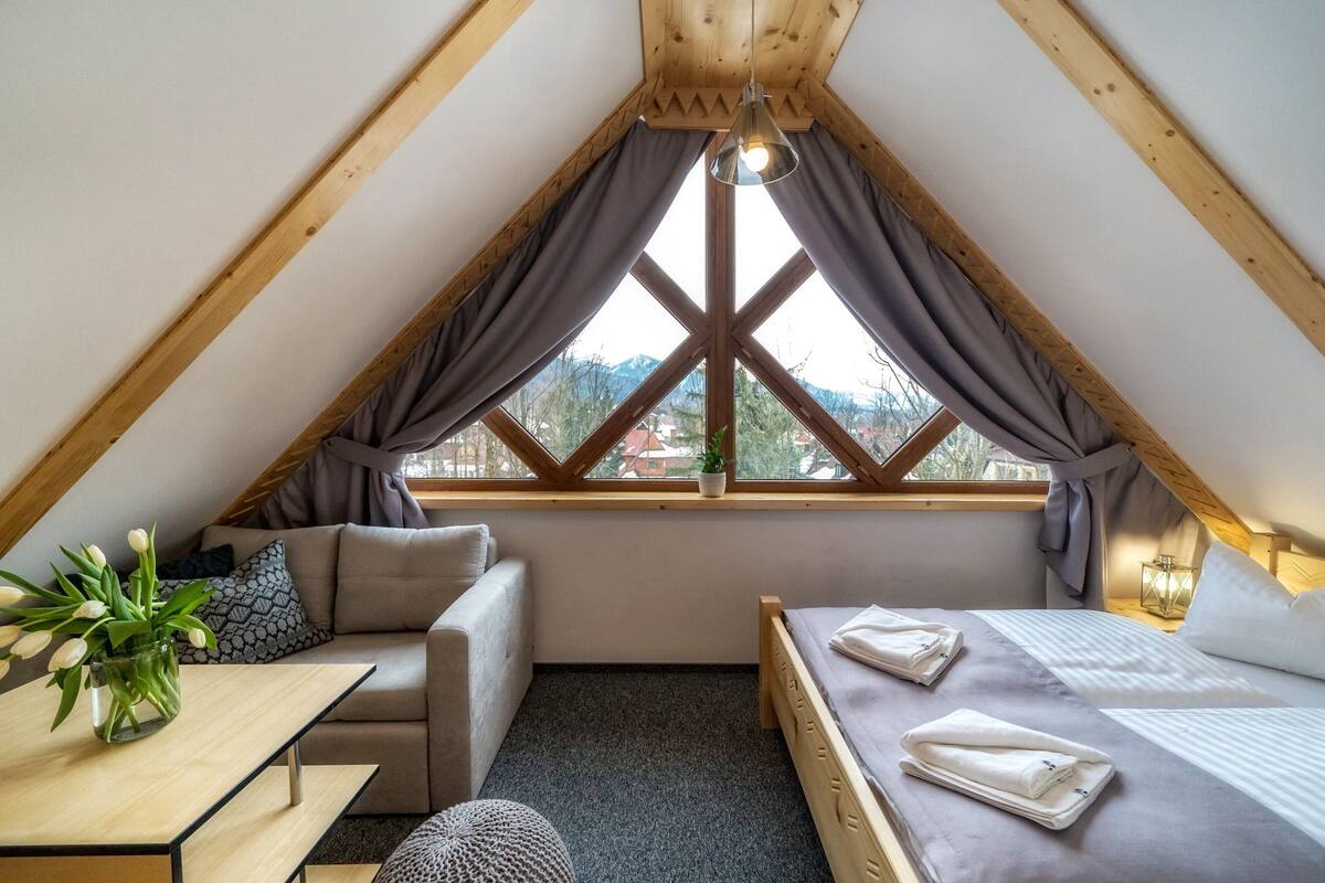A cozy attic bedroom with a bed, a sofa, a small table, and a window with curtains.