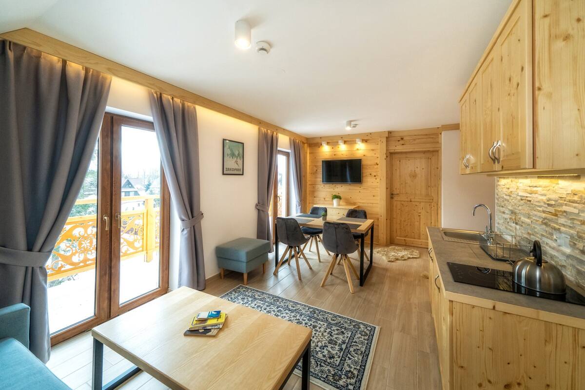 A compact room with a wooden dining table, a small kitchenette, and a large window with curtains.