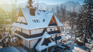 A multi-story chalet with a snow-covered roof, a balcony on the upper floor, and a parking area with several cars. - Kaplowa Chata (Zakopane)