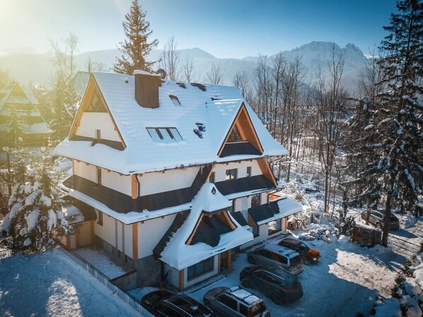 A multi-story chalet with a snow-covered roof, a balcony on the upper floor, and a parking area with several cars.