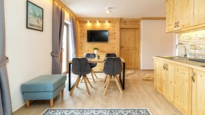 A compact kitchen with wooden cabinets, a dining table with four chairs, a small bench, and a flat-screen TV mounted on the wall. - Kaplowa Chata (Zakopane)