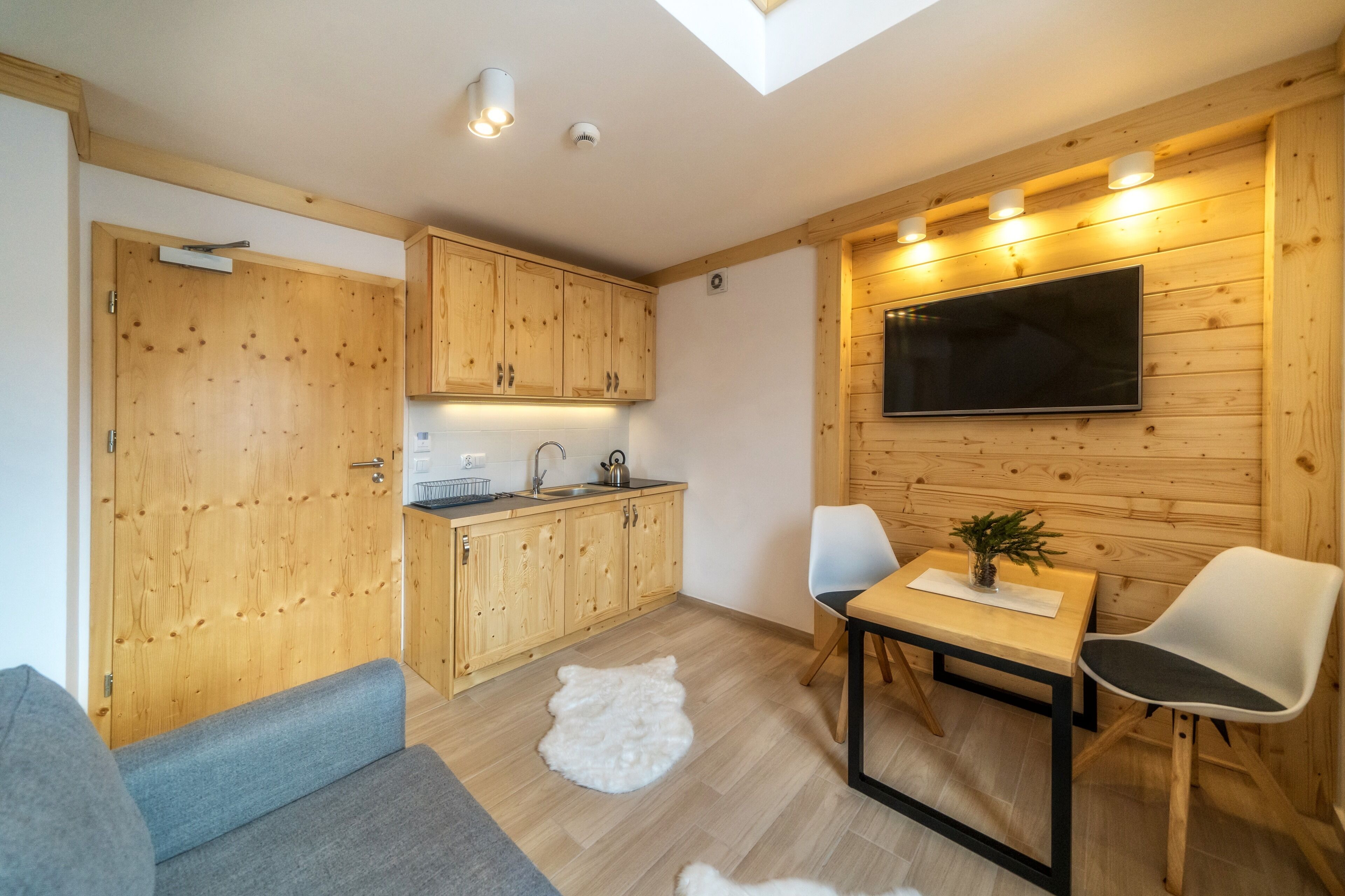 Un espacio habitable compacto con muebles de madera, un televisor de pantalla plana en la pared, una mesa de comedor con sillas y una zona de estar acogedora con un sofá y una alfombra blanca y mullida.