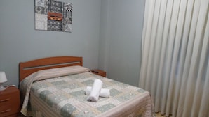 A bedroom with a wooden bed, a nightstand, a lamp, a quilt, and a window with curtains. - Pensión Cinco Villas (Logroño)