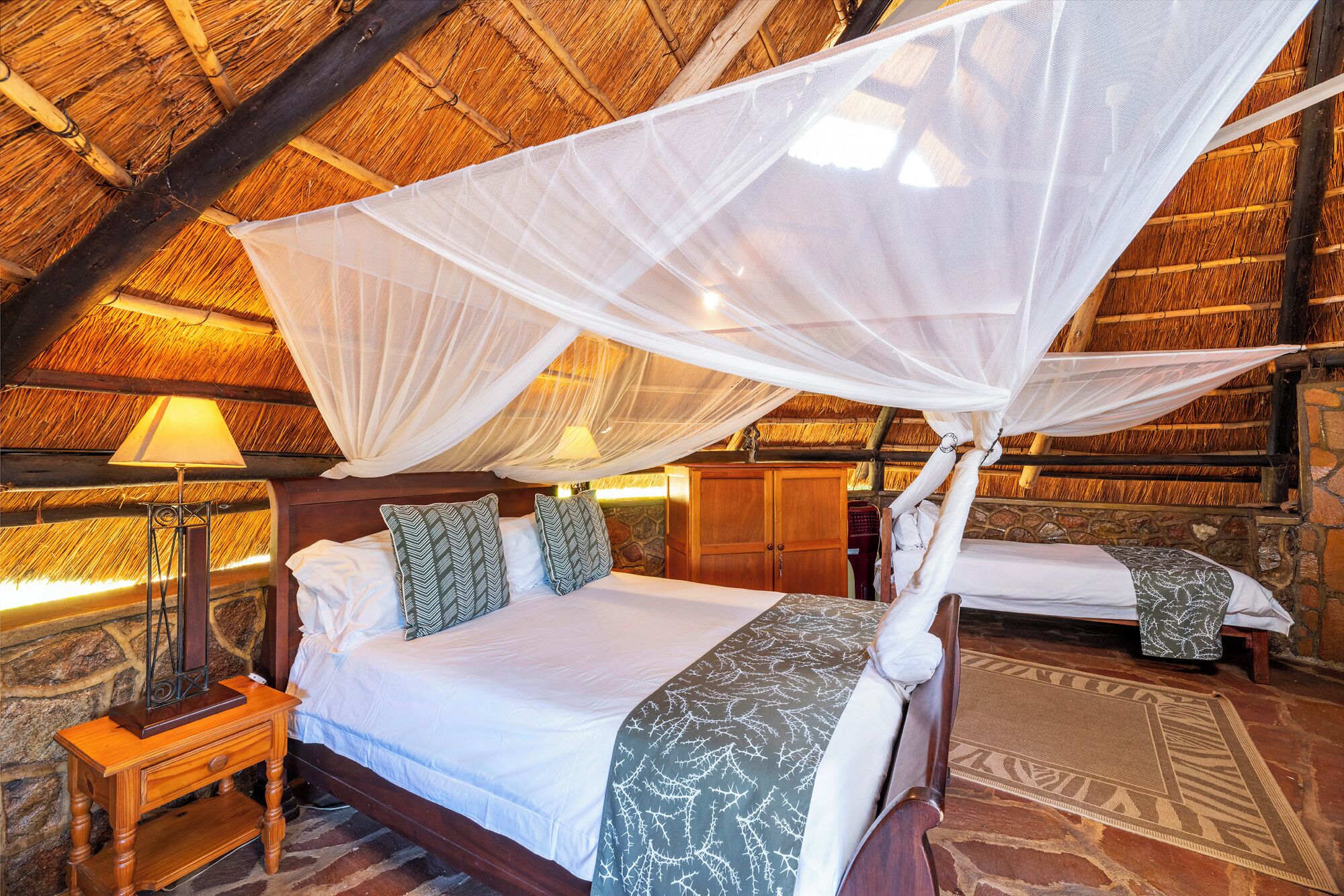 A room with a bed under a canopy, a nightstand with a lamp, and a stone wall.