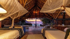 A thatched-roof bedroom with two beds, a patterned rug, and a view of the sunset over the ocean. - Hornbill Lodge (Kariba)
