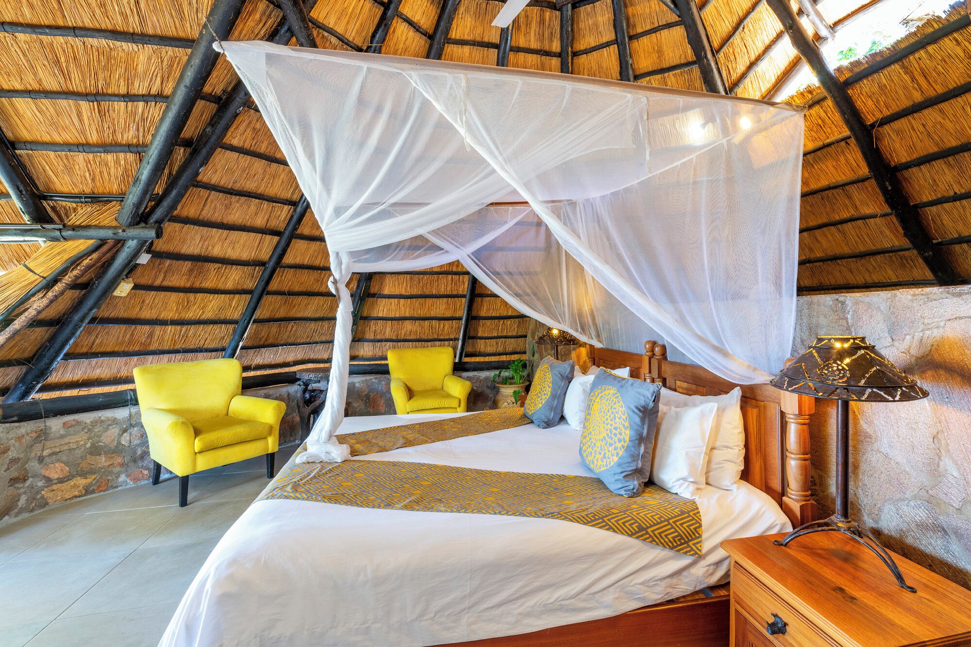Una habitación con una cama, sillones amarillos, una mesita de noche de madera y un mosquitero.