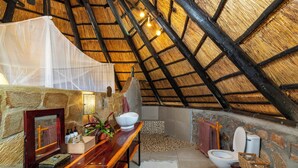 A rustic bathroom with a stone wall, wooden bench, and a thatched roof. - Hornbill Lodge (Kariba)