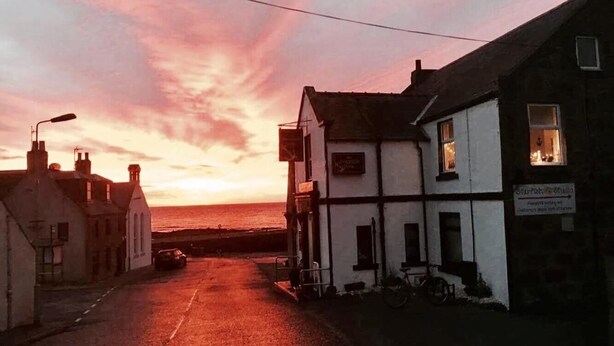 A coastal street at sunset with buildings on either side, a clear view of the sea, and a sign indicating a pub. - The Anchor Hotel (Montrose)