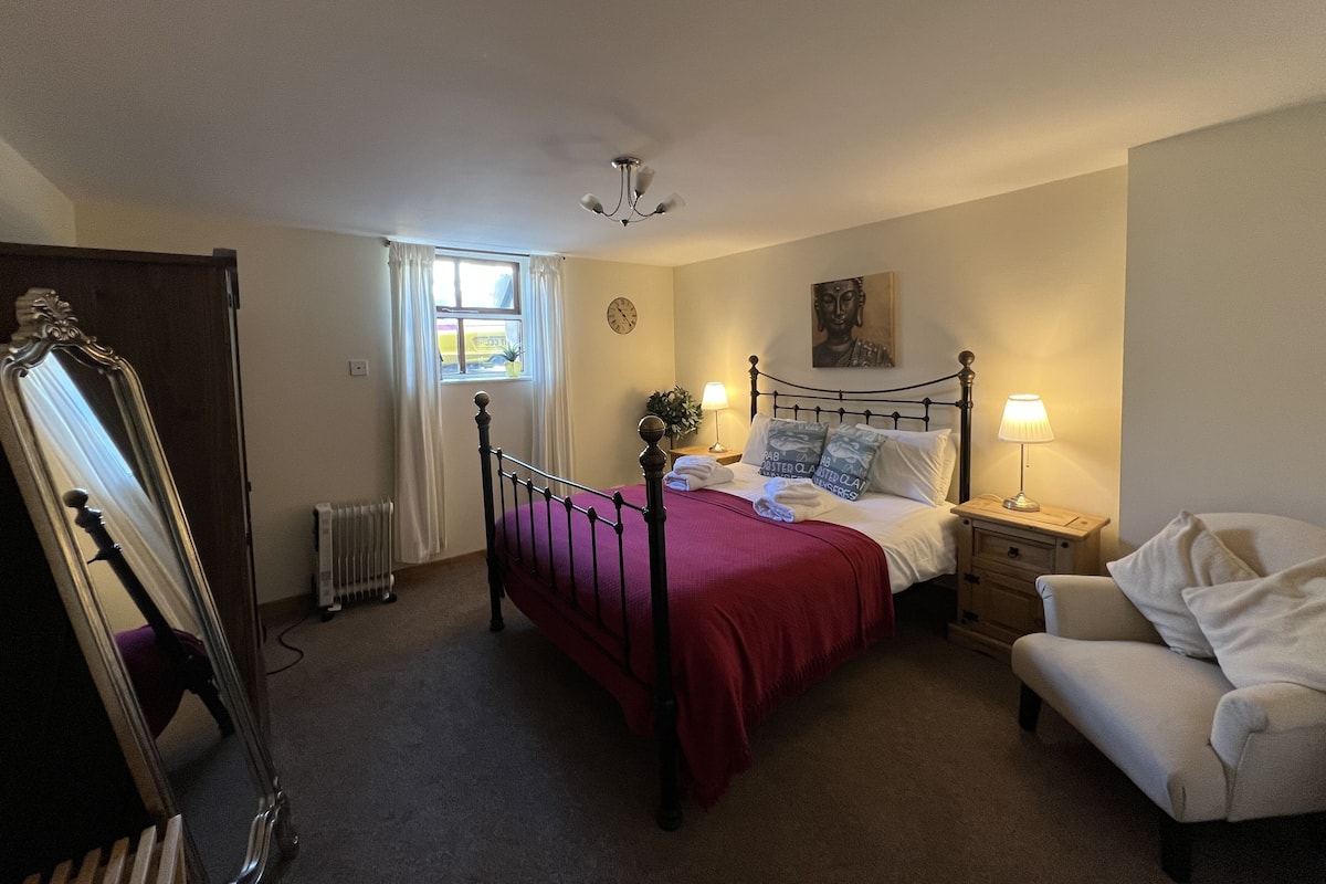 A bedroom with a bed, a chair, a bedside table with a lamp, a window with curtains, a radiator, and a mirror.