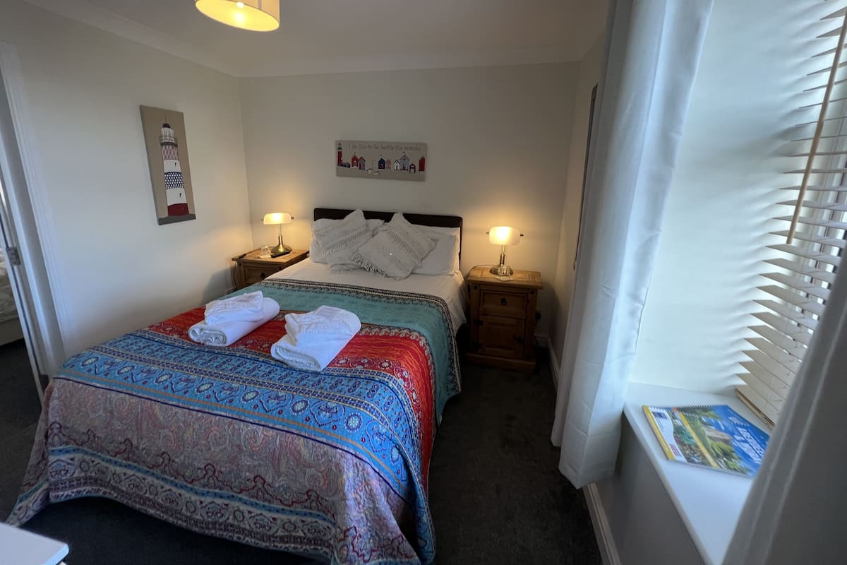 A neatly made bed with a patterned bedspread, two bedside lamps, a framed picture on the wall, and a window with blinds.