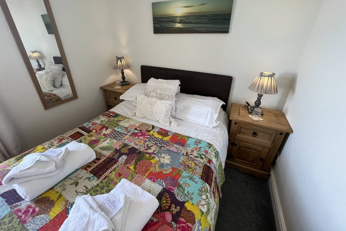 A neatly made bed with a colorful quilt, two bedside tables with lamps, a mirror, and a painting of a sunset.