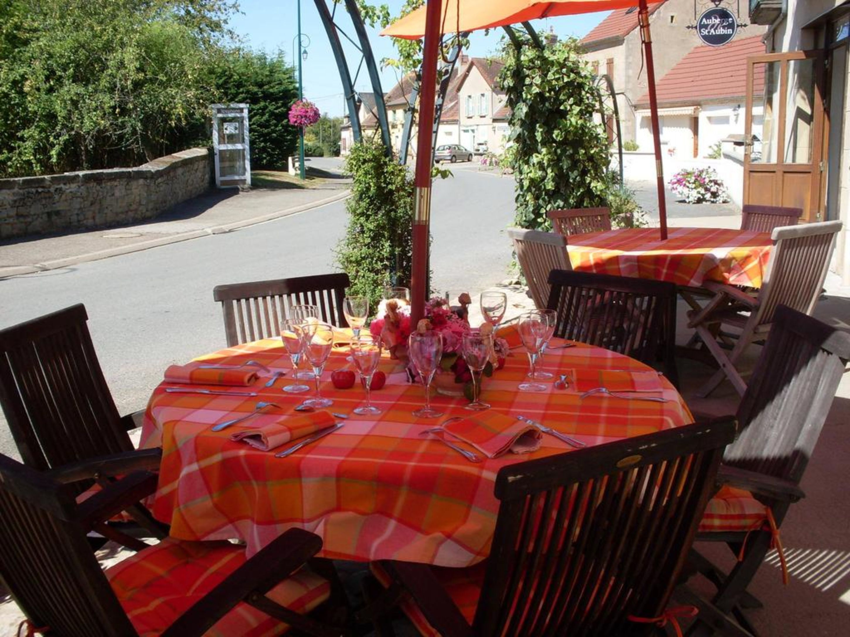 An outdoor dining area with round tables covered in orange and white checkered tablecloths, surrounded by wooden chairs.