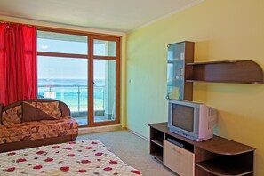 A bedroom with a bed, a sofa, a television on a wooden stand, and a large window with a view of the sea. - Black Sea Paradise (Sozopol)
