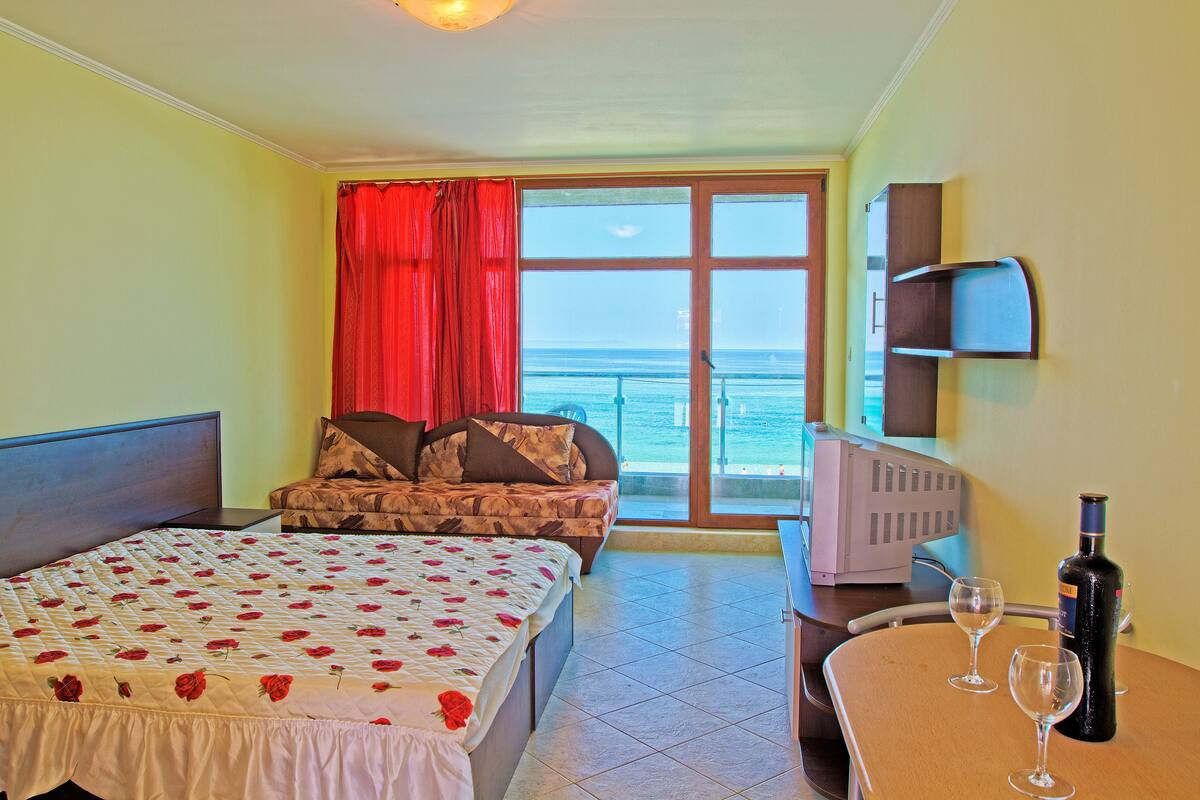 A hotel room with a bed, a sofa, a TV, a table with glasses, and a bottle.