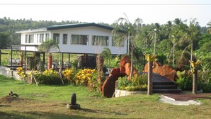 A building with a white exterior and green roof, surrounded by a landscaped garden with tropical plants and a large rock sculpture. - Vinewoods Resorts by Cocotel (Taal)