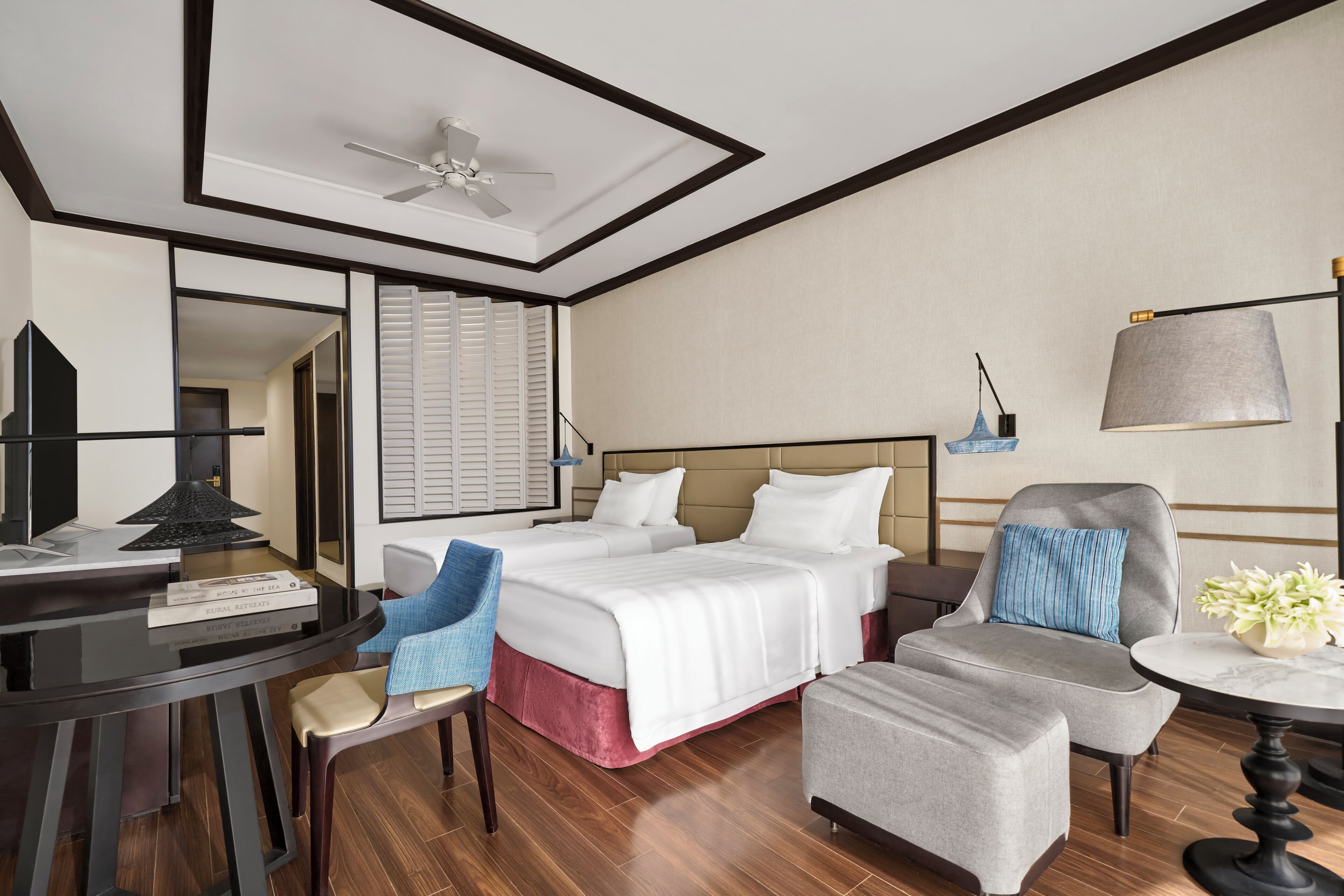 Een hotelkamer met een bed, bureau, stoel, tafel, lamp en plafondventilator.