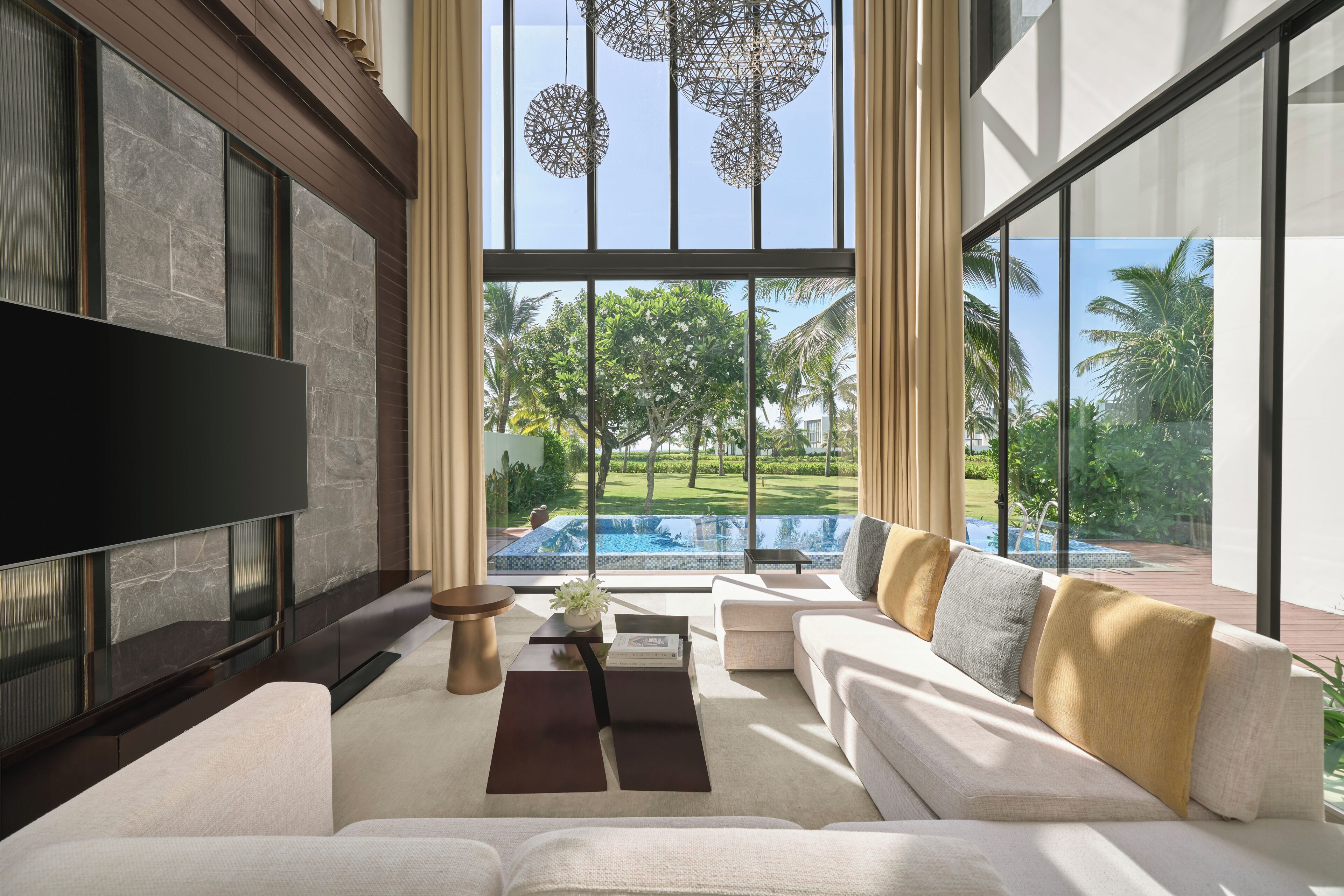 A modern living room with a large flat-screen TV, a sofa, a coffee table, and a view of a swimming pool and greenery.