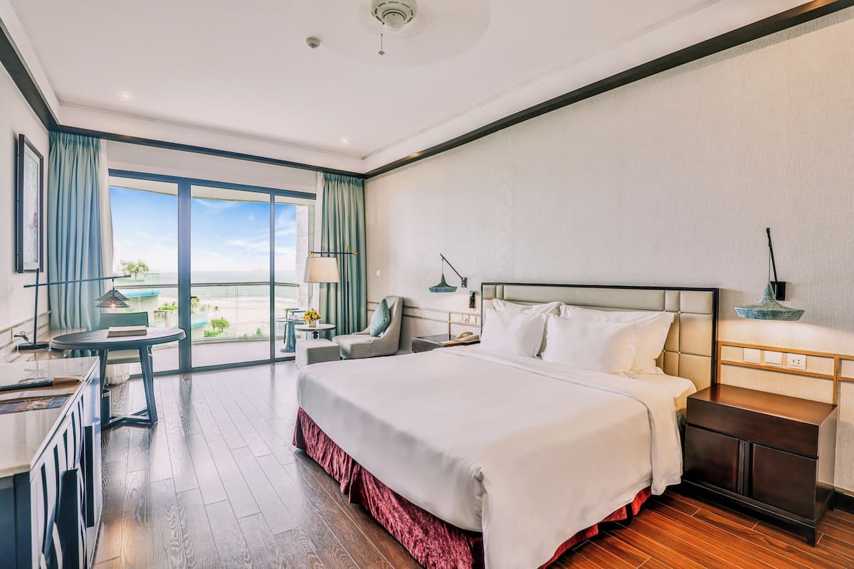 Une chambre d’hôtel avec un grand lit, un bureau, une chaise, une petite table avec une lampe et une vue sur l’extérieur.