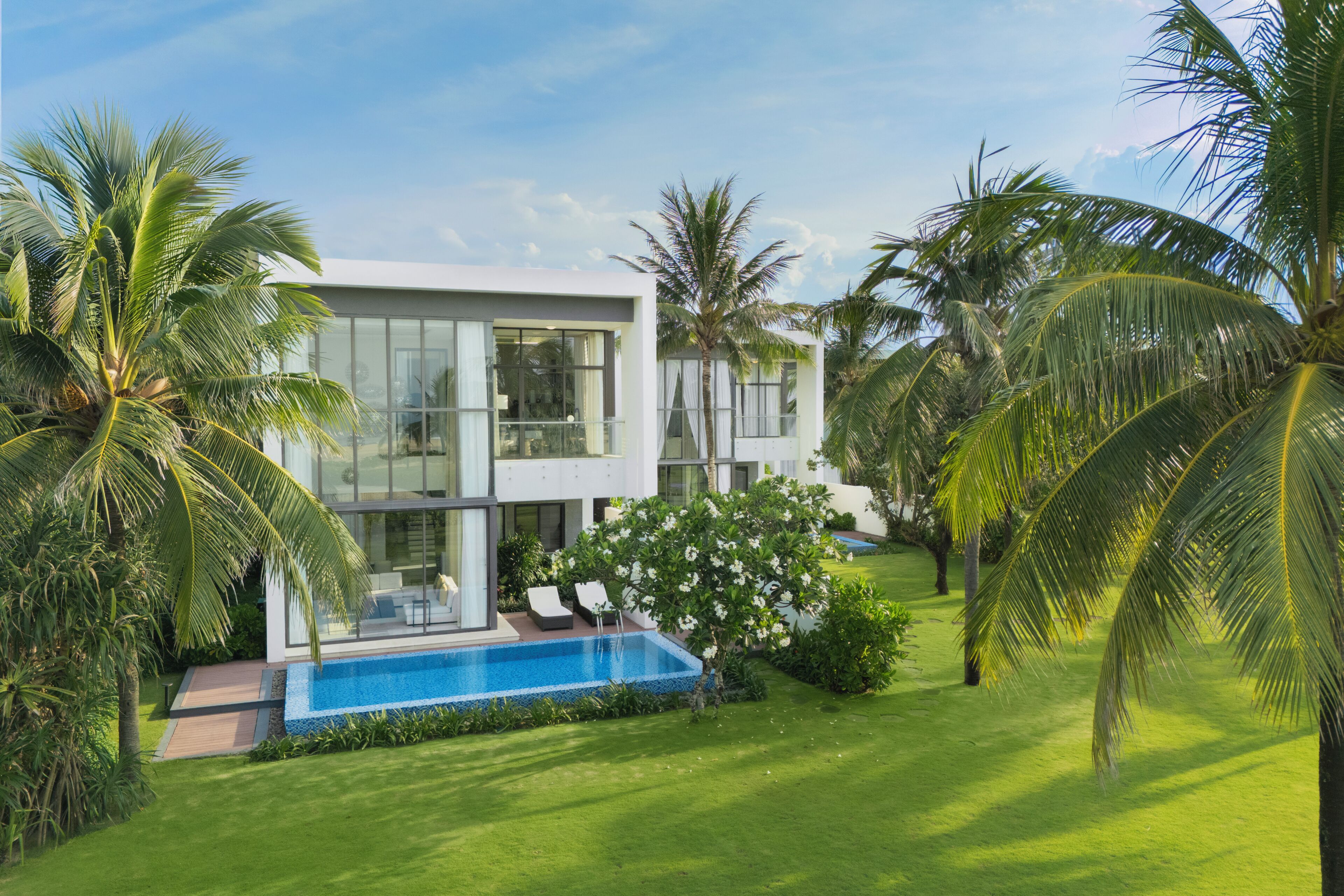 Uma casa moderna de dois andares com piscina, cercada por vegetação exuberante e palmeiras.