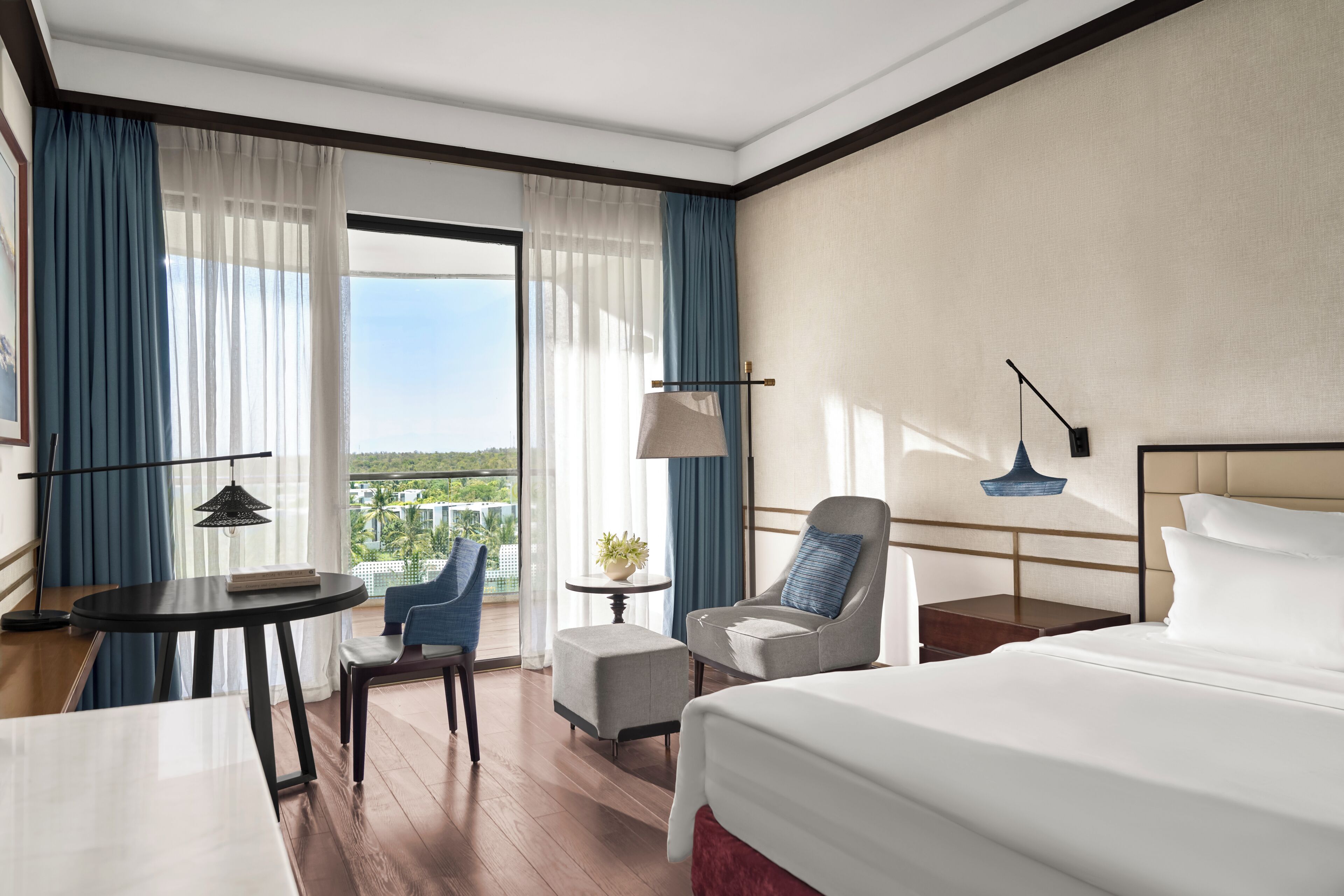 A hotel room with a bed, a desk, a chair, a small table with a plant, and a view of the outdoors.
