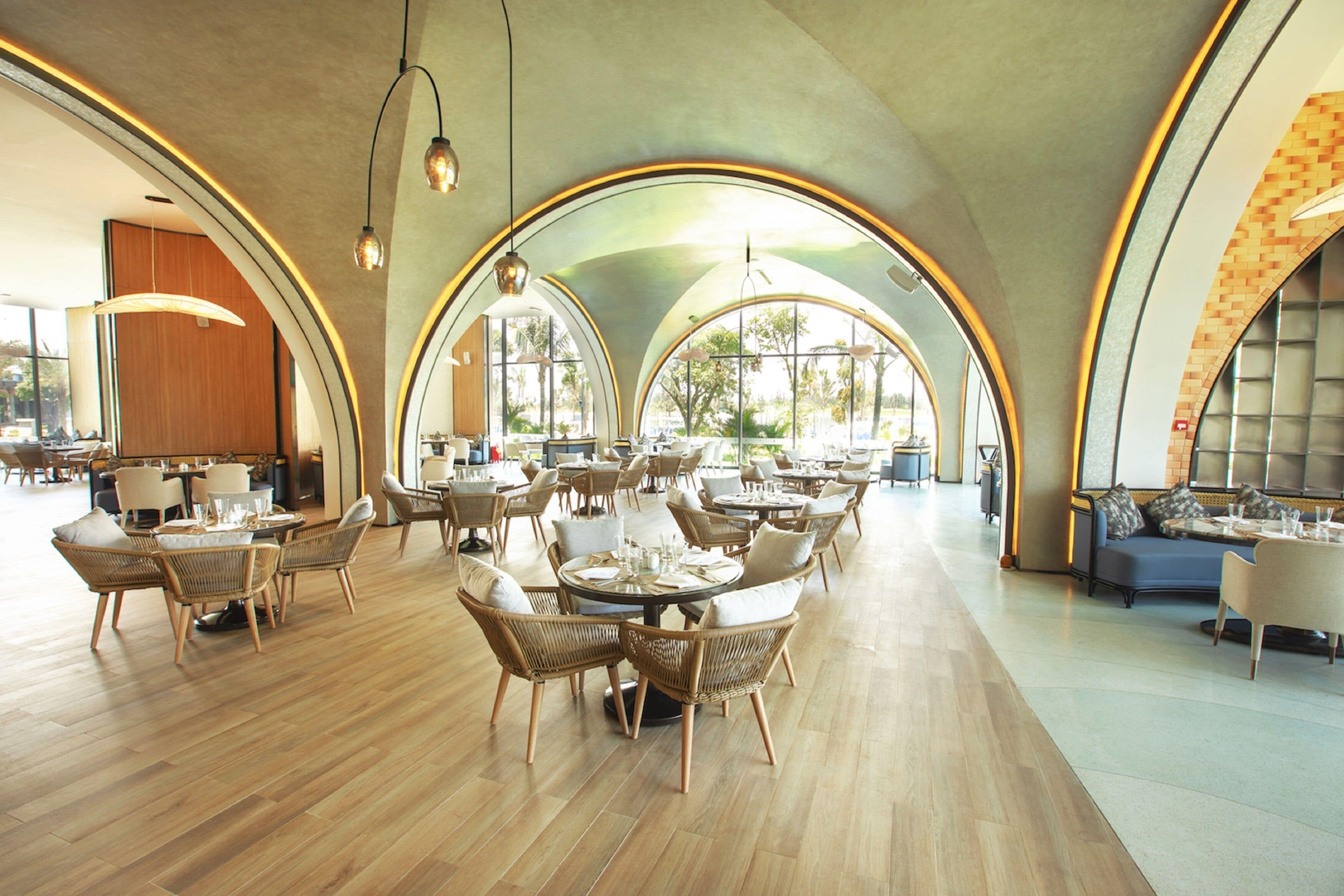 A spacious dining area with high arched ceilings and natural light from large windows. Tables are set with chairs, and the floor is wooden.