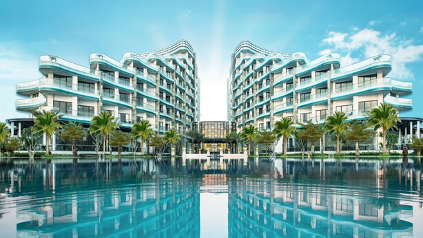 Un hôtel à l’architecture moderne, de conception symétrique, avec une piscine centrale dont l’eau reflète la lumière et des palmiers.