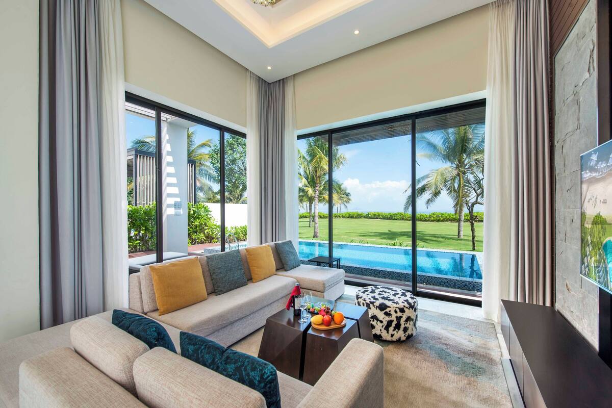 Un salon moderne avec un canapé, une table basse et un téléviseur. De grandes fenêtres offrent une vue sur une piscine et des palmiers.