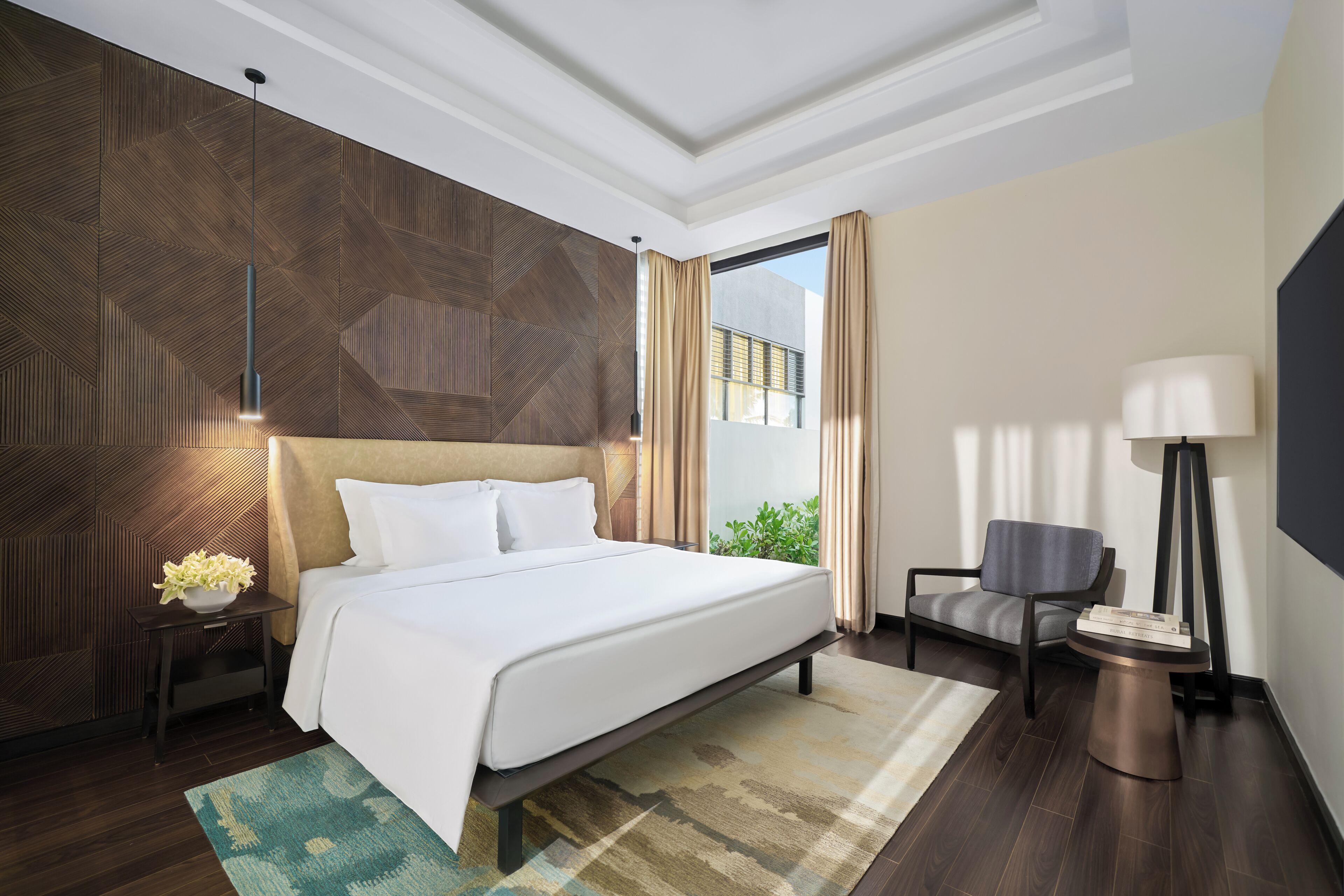 Quarto de hotel moderno com uma cama grande, mesa de cabeceira com abajur, uma poltrona e vista para o verde lá fora.