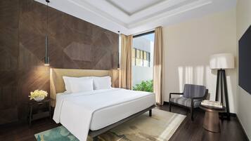 Quarto de hotel moderno com uma cama grande, mesa de cabeceira com abajur, uma poltrona e vista para o verde lá fora.