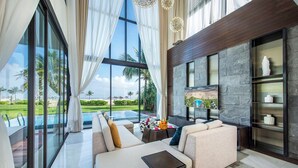 Un salon moderne avec un grand canapé, une table basse et un téléviseur. Il y a une grande fenêtre avec des rideaux, un mur en pierre et une vue sur une piscine et la verdure environnante.