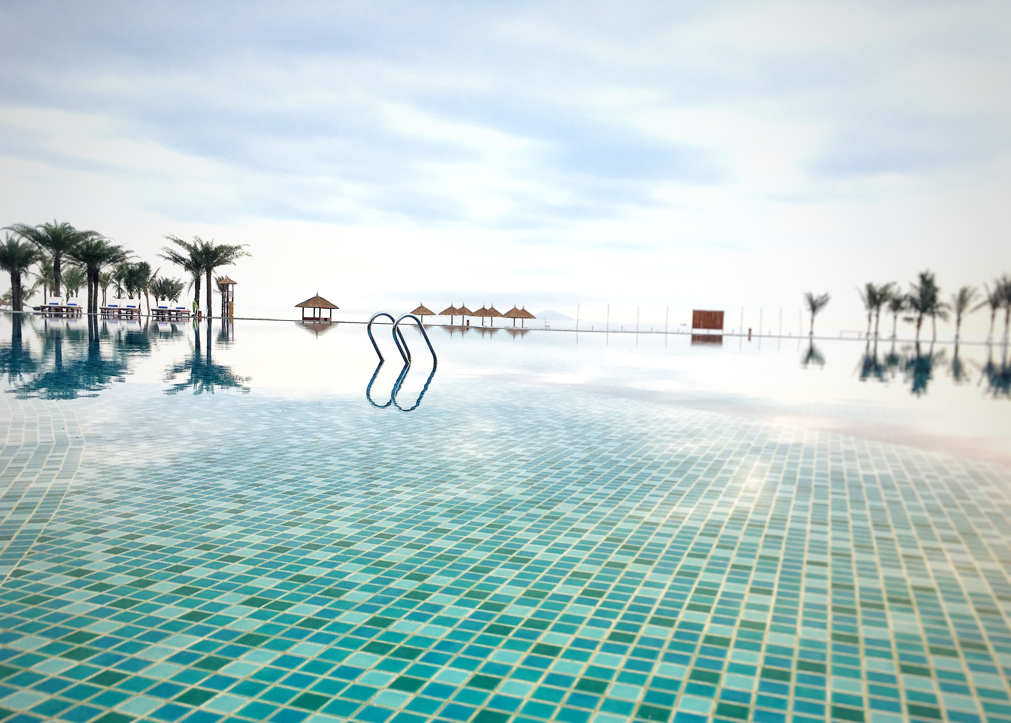 An infinity pool with clear blue tiles, palm trees, and beach huts in the background.